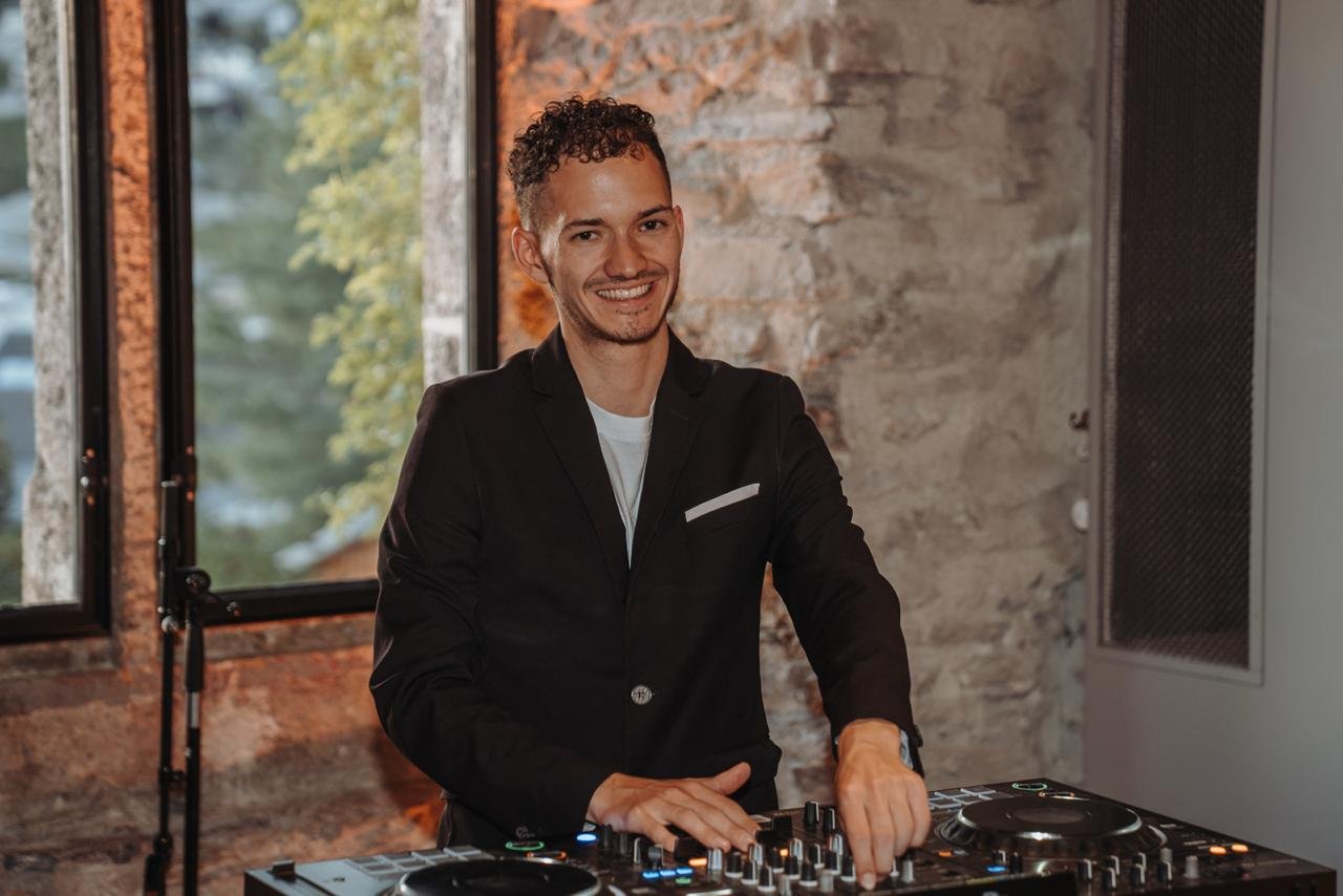 Cale lächelt im dunklen Sakko hinter dem DJ-Pult vor einer Steinwand